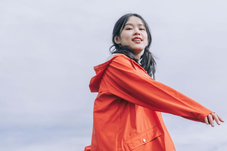 Mustatukkainen tyttö oranssissa sadetakissa, taustalla sinertävä taivas. 