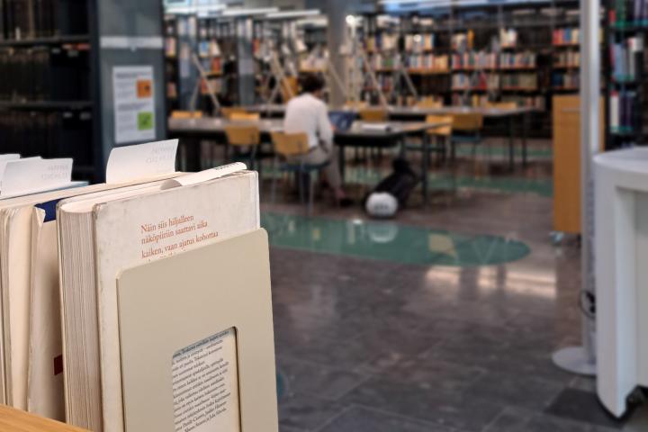 Etualalla kirjoja hyllyssä, taustalla henkilö opiskelee kirjahyllyjen välissä.