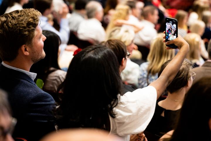 Juhlayleisössä istuva henkilö ottaa selfie-kuvaa.